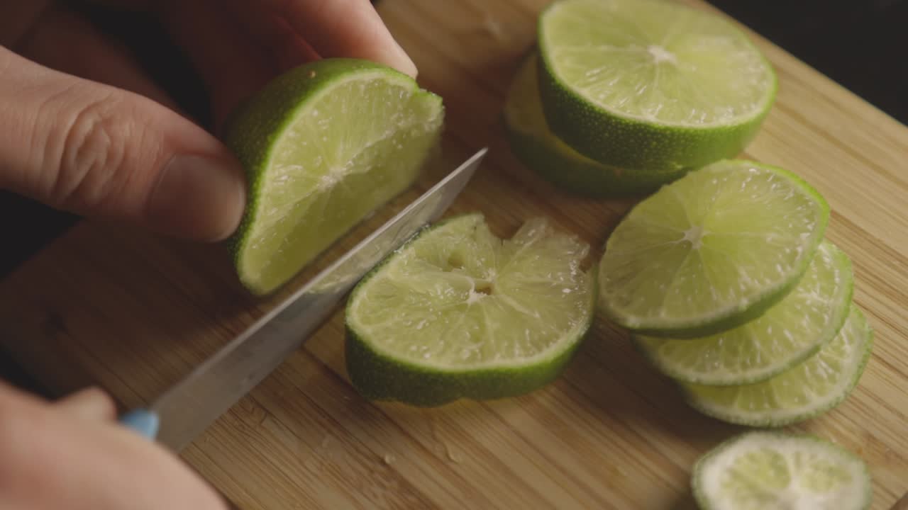 Hand Firmly Holding A Lime Fruit While Cutting It Into Thin Slices Using A Sharp Kitchen Knife - Closeup Shot