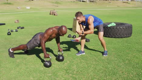 two diverse fit men exercising outdoors, one doing push ups on kettlebells while the other times him