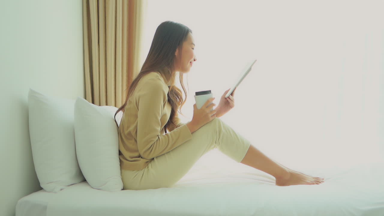 A young woman propped up with pillows sips coffee while reading a book