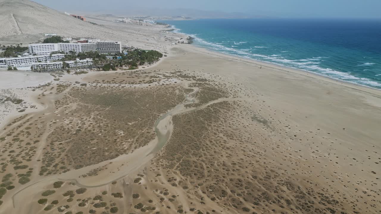 Drone footage of Sotavento Beach in Fuerteventura, showing ocean waves, golden sand, and a hazy sky. Ideal for travel, nature, and coastal lifestyle themes.