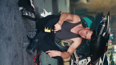 Vertical-video:-a-male-mechanic-with-a-short-haircut-in-a-gray-shirt-sits-on-the-floor-drinking-a-drink-from-a-brown-glass-bottle-in-a-workshop-garage