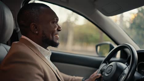 Un-Hombre-De-Negocios-Feliz-Y-Sonriente,-De-Piel-Negra-Y-Barba-Con-Una-Chaqueta-Marrón,-Conduce-Un-Automóvil-Moderno-Durante-Su-Viaje-De-Negocios-Por-La-Ciudad