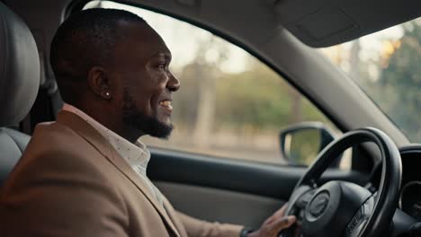 happy and smiling man businessman with black skin color and beard in a brown jacket drives a modern car during his business trip around the city