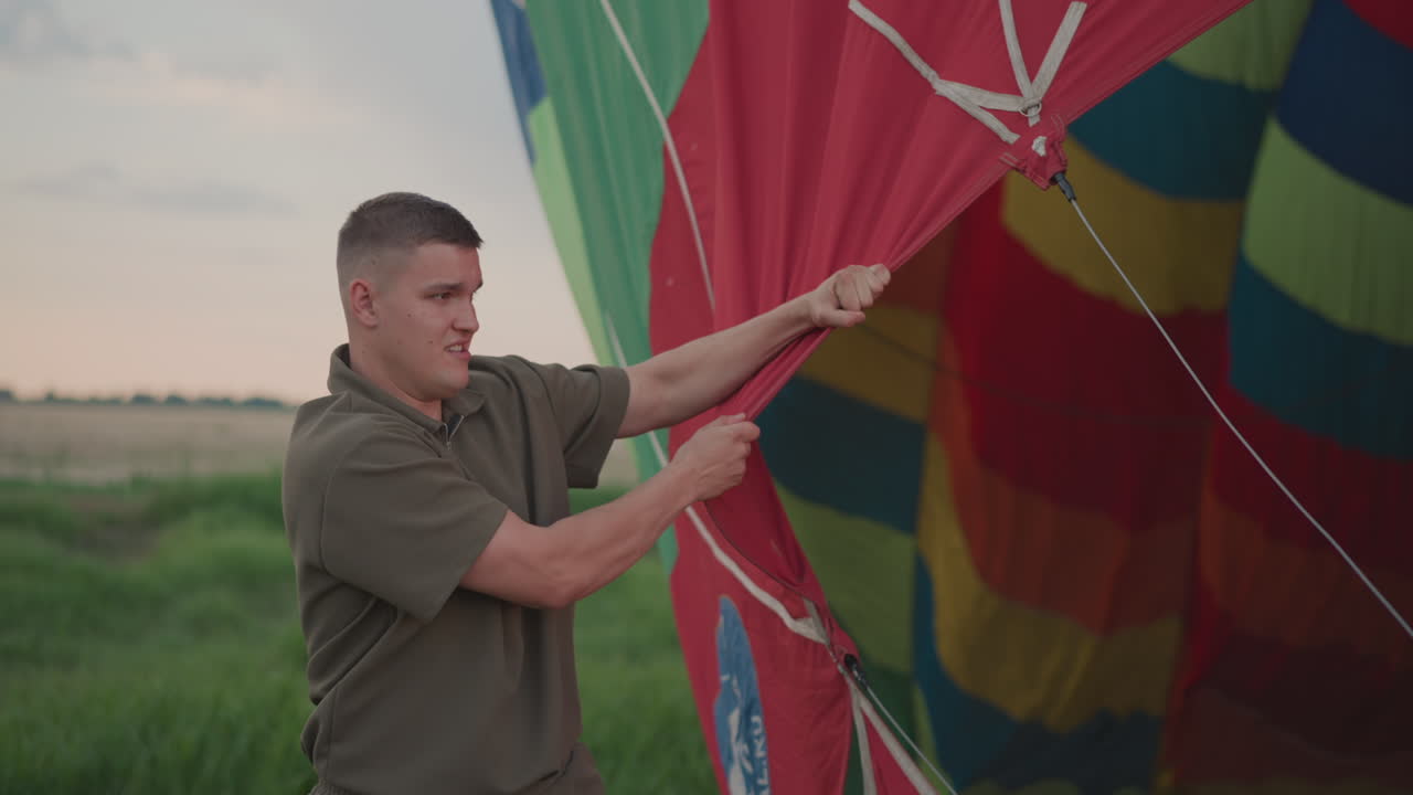 man gripping hot air balloon envelope material during preflight setup in vast open field at dusk pulling tensioned ropes to hold envelope open for burner inflation next to basket and engine fan