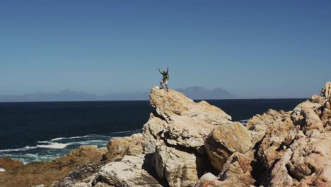 African-american-man-hiking-raising-hands-standing-on-rock-by-the-sea