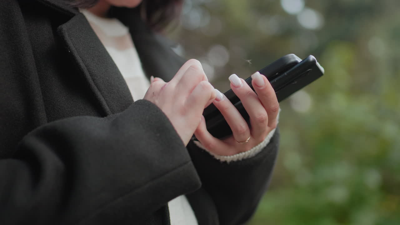 가을을 사랑하는 사람이 핸드폰 두 대를 들고 손톱이 예쁘게 관리된 손으로 위쪽 화면을 터치하며 밝은 표정을 짓고 있어요. 공원에서 아늑한 코트를 입고 있으며 배경은 부드러운 보케로 처리되어 있고, 현대적인 소통의 순간을 담고 있습니다.