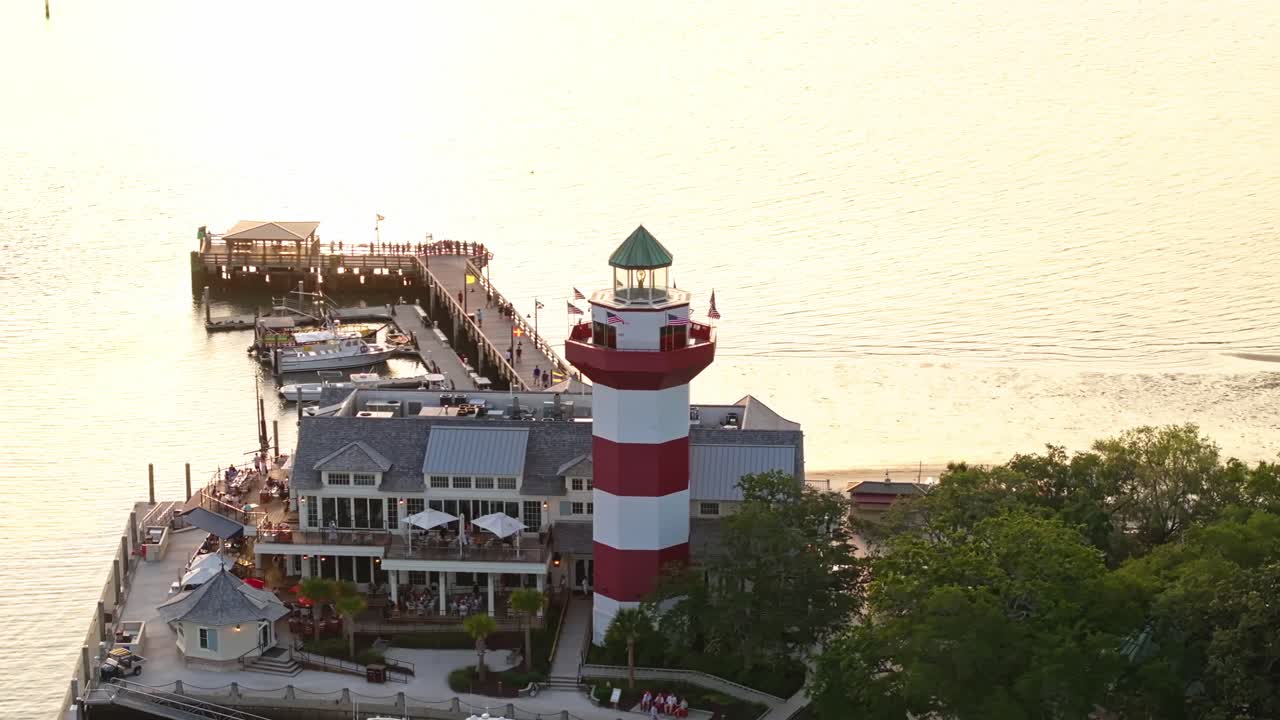Lighthouse reflecting in harbor water during vibrant sunset over pier, telephoto aerial orbit, Harbour Town Pier South Carolina USA