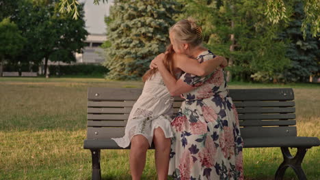 a young woman confides in her mother about her day. they sit on a park bench and share a tender hug. dolly shot to the right in warm natural light