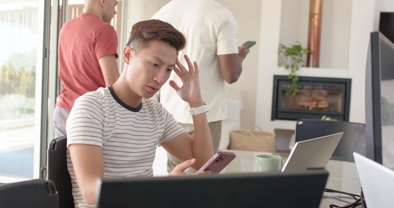 Diverse men touching shoulder while Asian man sitting at table reacting checking pink phone laptop