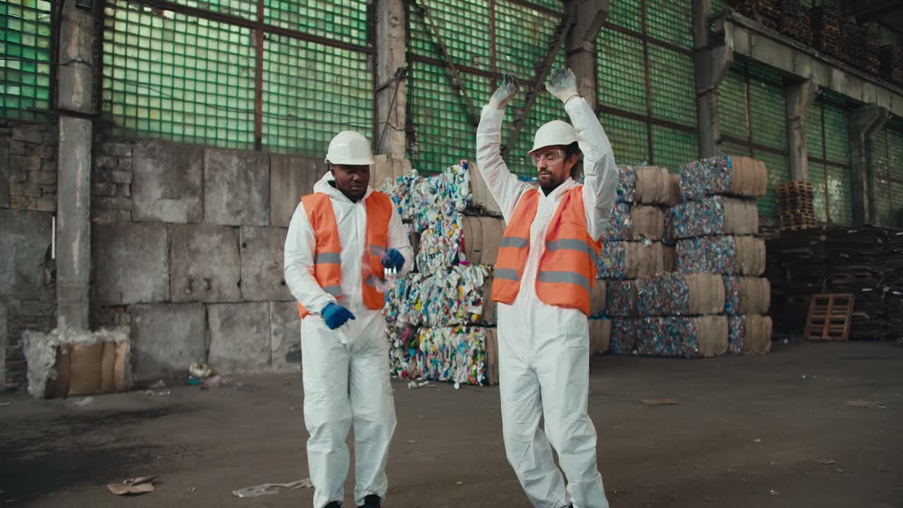 Approaching two happy male colleagues, a man with Black skin in a white protective uniform in an orange vest, together with his employees a man with a beard and in a white protective helmet dances near the racks and sorted garbage in the large hall of a waste sorting and recycling plant