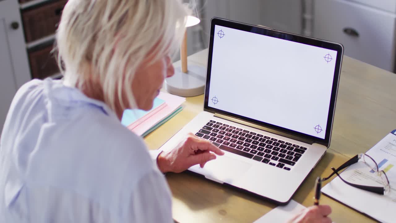 mujer caucásica mayor feliz sentada en la mesa en la cocina, usando una computadora portátil con espacio de copia