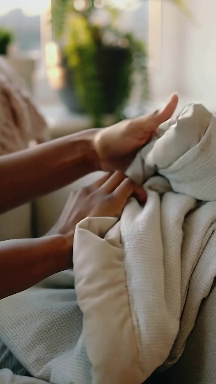 Video verticale: Mani femminili che piegano una coperta sul divano vicino alla finestra, sistemando un cuscino per riordinare.