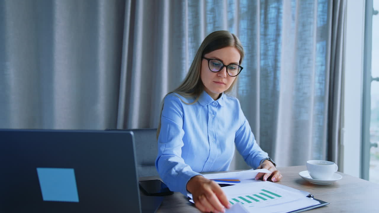 Busy lady wearing glasses working in office at her desk. Employee looks through the documents quickly, turning pages. Grey backdrop.