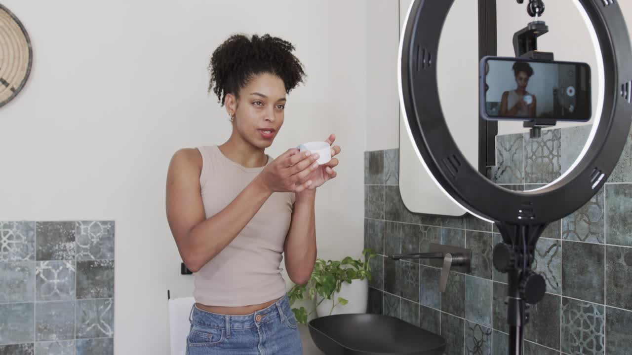 Adult African American woman leaning at vanity, picking jar, scooping product with spatula to demo