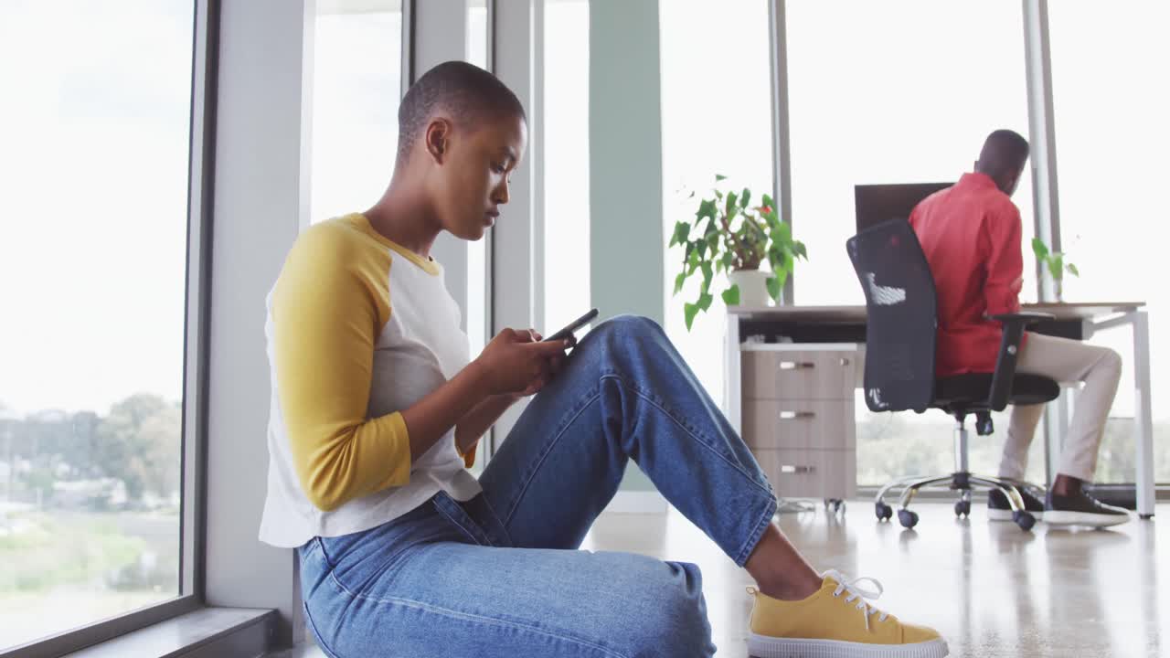 African american creative businesswoman sitting on floor using smartphone, male colleague at desk