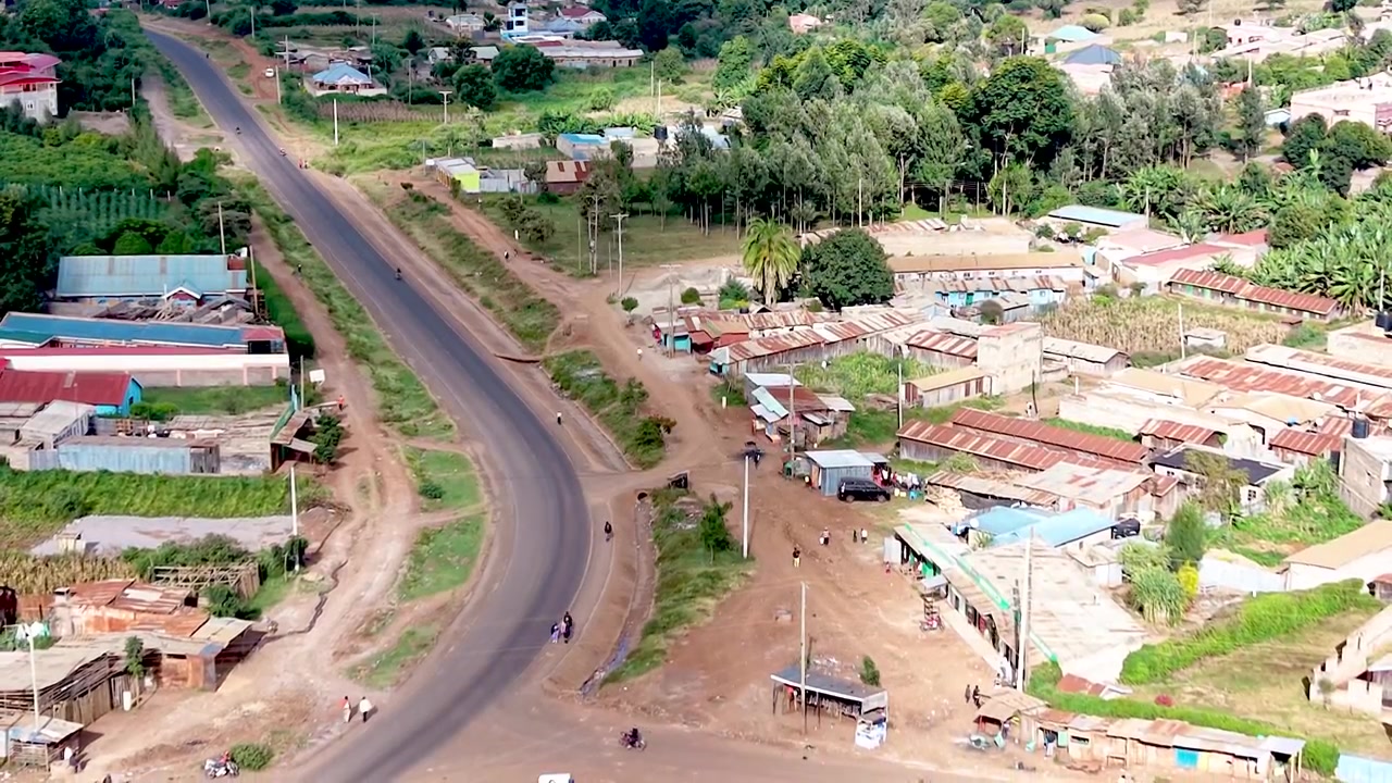 Una vista cinematografica del Kenya rurale che mostra insediamenti informali con tetti di lamiera arrugginiti, case logore, sentieri polverosi del villaggio e la vita quotidiana della comunità.