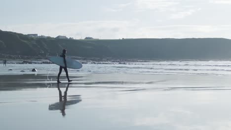 Tiro-Medio-Ancho-De-Un-Surfista-En-Un-Traje-Húmedo-Que-Lleva-Su-Tabla-De-Surf-Bajo-El-Brazo-En-Una-Playa-Húmeda-En-Irlanda