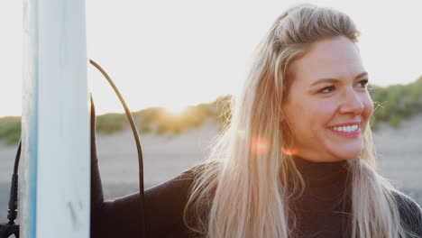 Mujer-Vistiendo-Traje-De-Neopreno-Sosteniendo-Una-Tabla-De-Surf-Disfrutando-De-Unas-Vacaciones-De-Surf-En-La-Playa-Mientras-Se-Pone-El-Sol