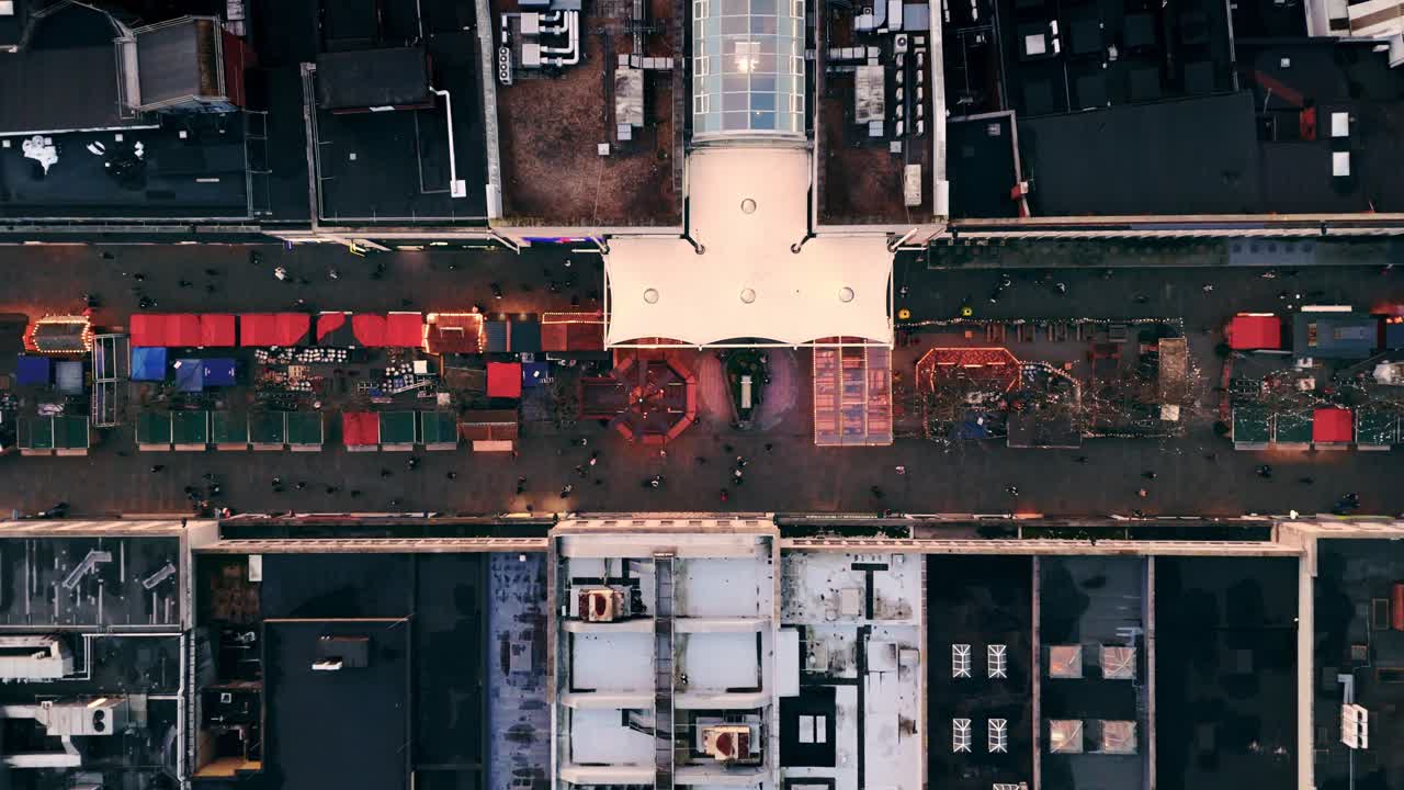Bird’s-eye drone shot rising over Southampton’s Christmas Market during a winter sunset, revealing glowing festive stalls, colorful lights, and crowds exploring as the city softens under warm light
