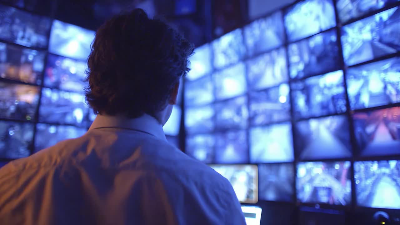 A Focused Individual Analyzes Surveillance Footage in a Dark Monitoring Room Surrounded by Multiple Screens Displaying Live Video Feeds from Various Cameras