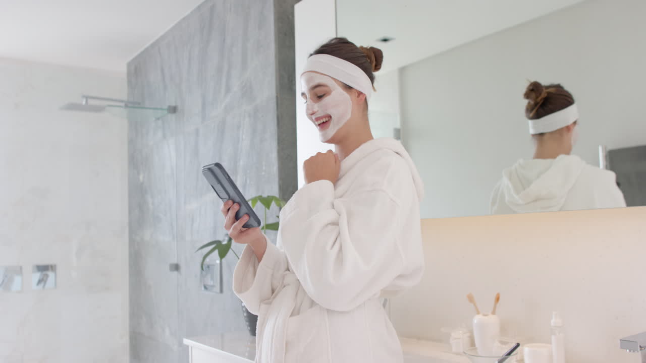 Applying face mask, woman in bathrobe using smartphone in bathroom, at home