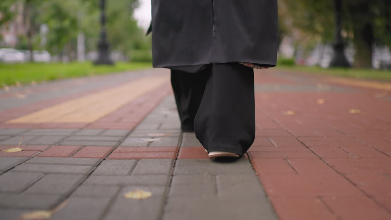 Close-up of young woman walking on park pathway in black coat and sneakers, one foot lifted mid-step, autumn leaves scattered on ground, evoking sense of motion, solitude, freedom, seasonal change