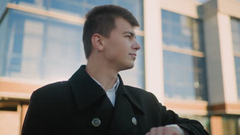 man in black clothing checks wristwatch while gazing into distance, exuding confidence and determination, background featuring modern glass building illuminated by soft natural light