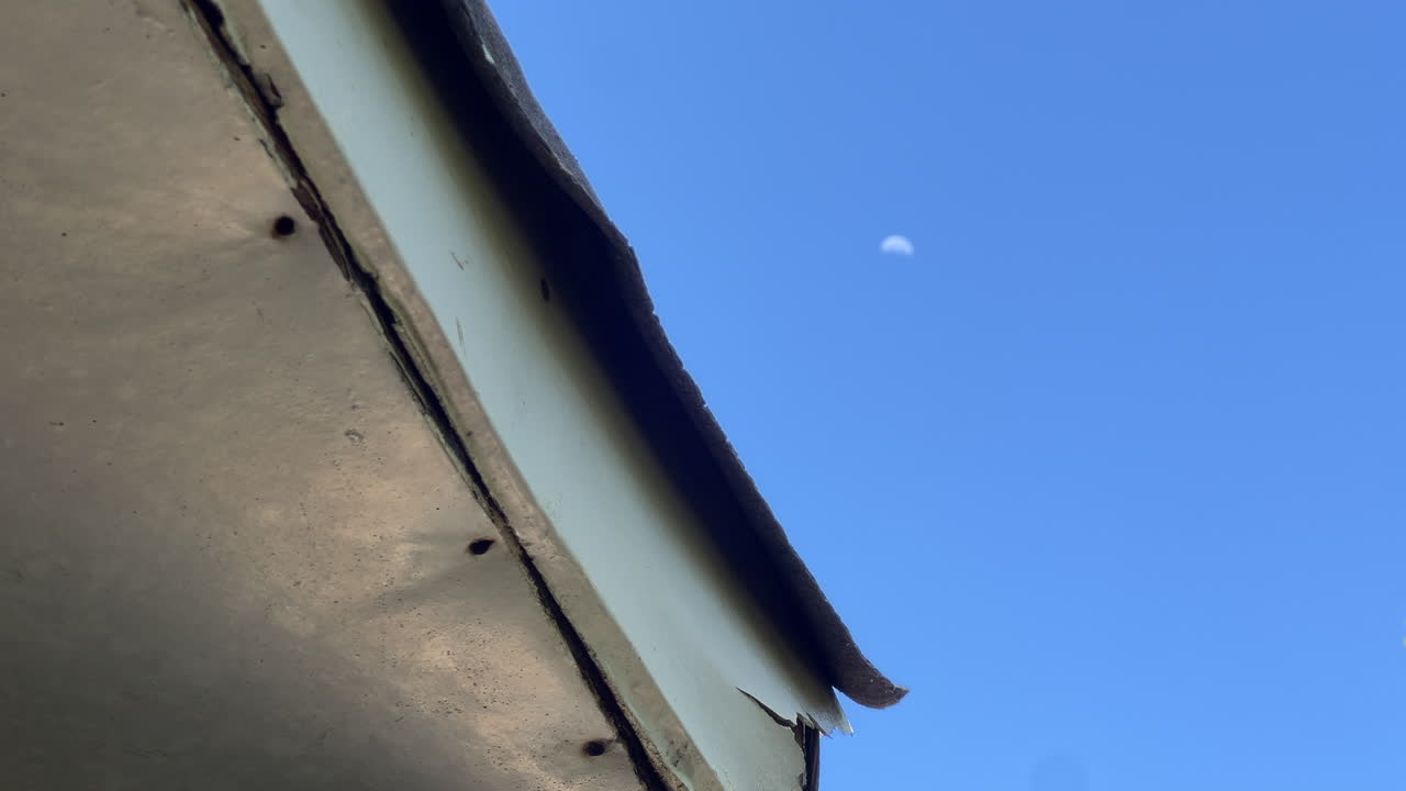 Damaged roof eave against clear blue sky with soft moon bokeh, highlighting neglected maintenance and repair needs