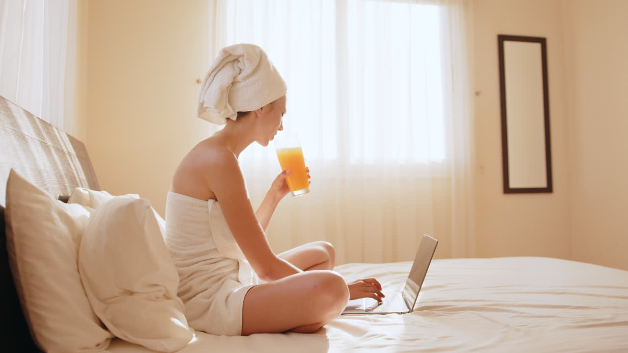 오렌지 주스를 마시면서 침대에서 노트북을 쓰고 있는 여자