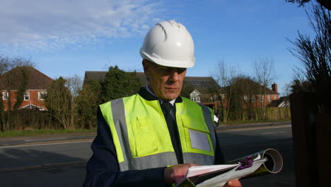 Static-shot-of-an-architect-senior-building-construction-manager-on-a-residential-street-with-traffic-and-houses-looking-at-paperwork-inspecting-the-building