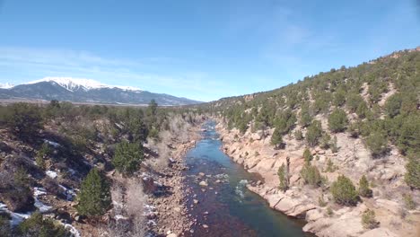 Drone-view-of-a-green-river-in-Colorado-rocky-ground-with-snowy-mountains-in-the-background