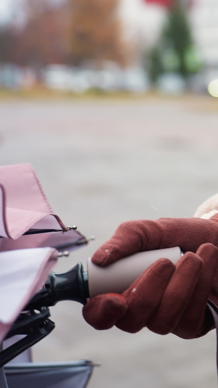 Close-up of gloved hand holding umbrella handle, person wearing brown shearling jacket with zip, standing outdoors on cold day. Face not visible, soft background blur and winter atmosphere