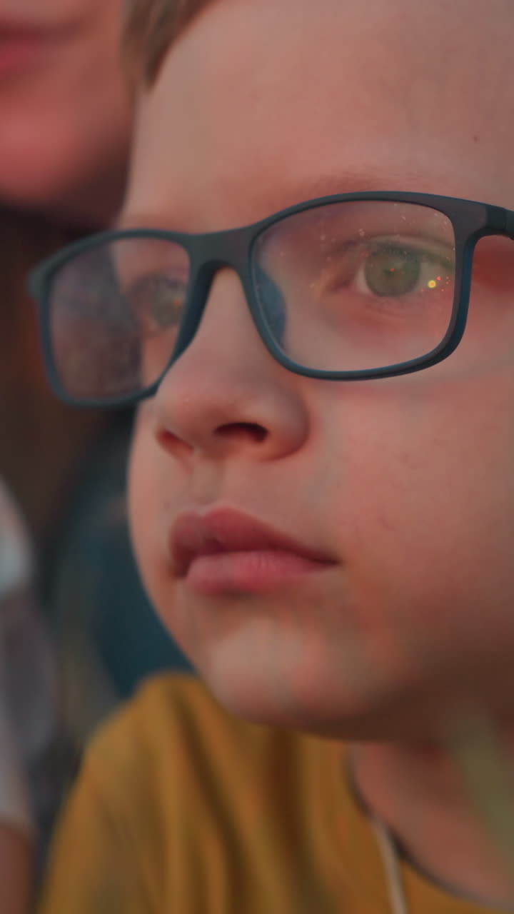 una toma de primer plano de un niño que lleva una camisa amarilla y gafas, profundamente enfocado en lo que está observando. en el fondo, una mujer y un niño más joven están suavemente borrosos