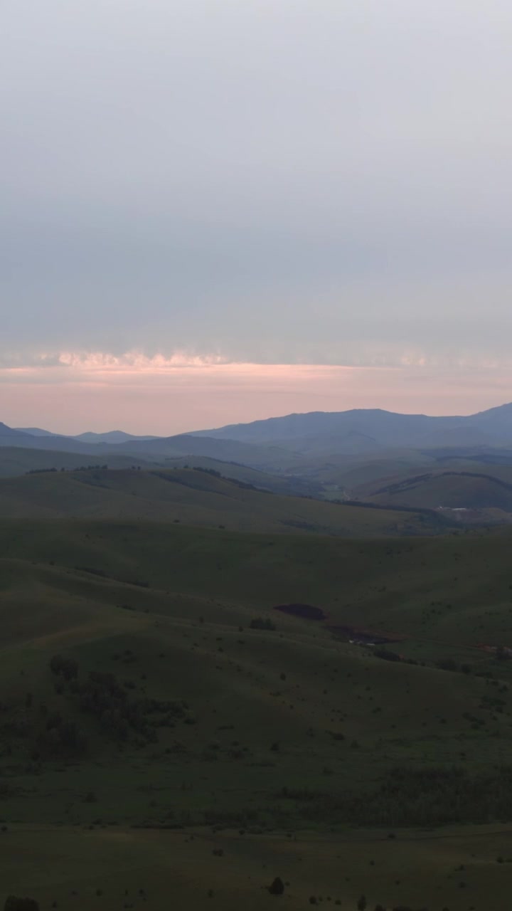 Wide landscape footage of rolling green hills and distant mountain ridges at sunset in the Altai region, Russia, with soft pastel light and dramatic overcast clouds