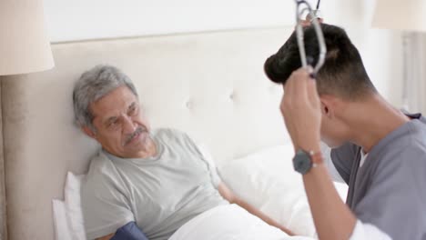 diverse male doctor testing blood pressure of senior male patient in bed at home, slow motion