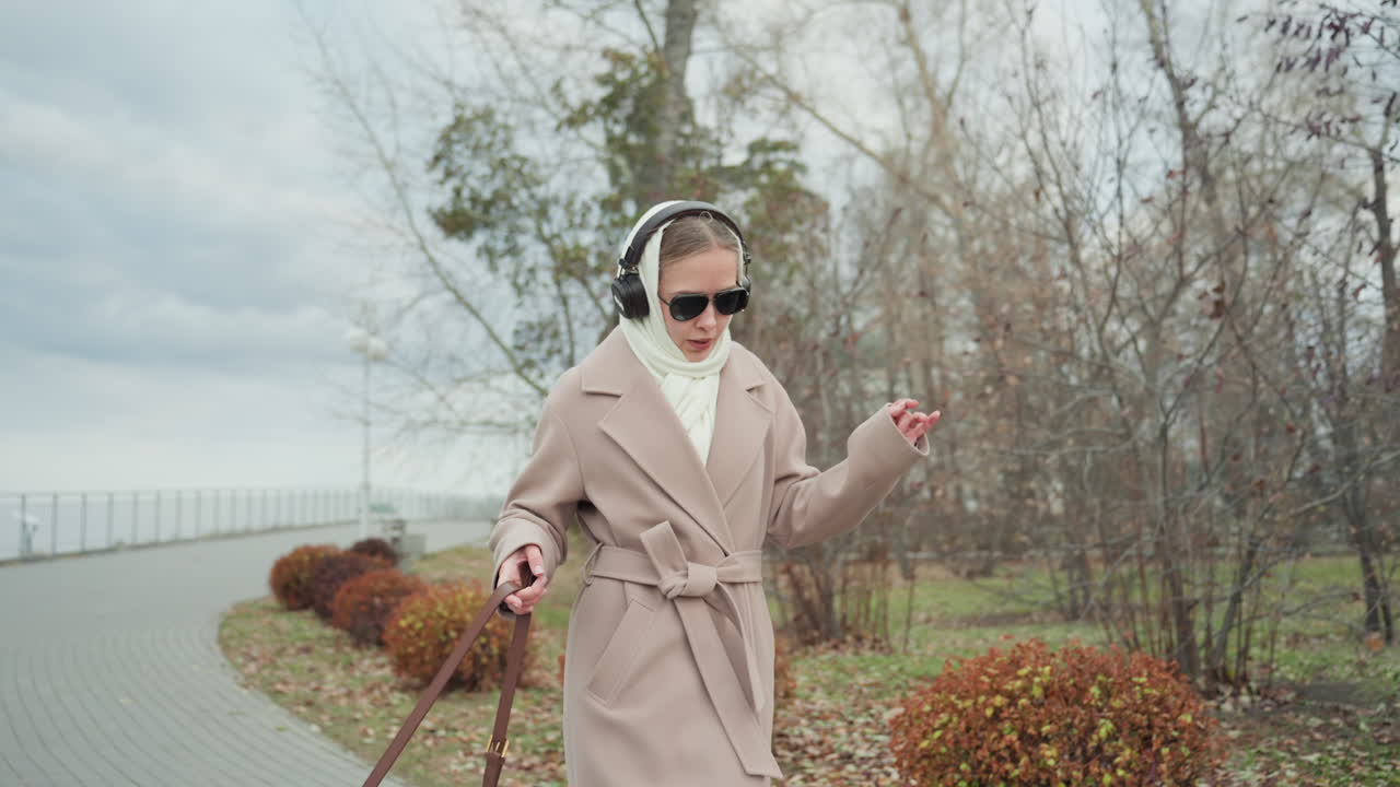 Donna caucasica serena che passeggia lungo la passeggiata in cappotto beige e cuffie, occhiali da sole che riparano lo sguardo, passeggiata lenta oltre alberi spogli e siepi potate, cielo coperto, lieve sorriso, mani in equilibrio.