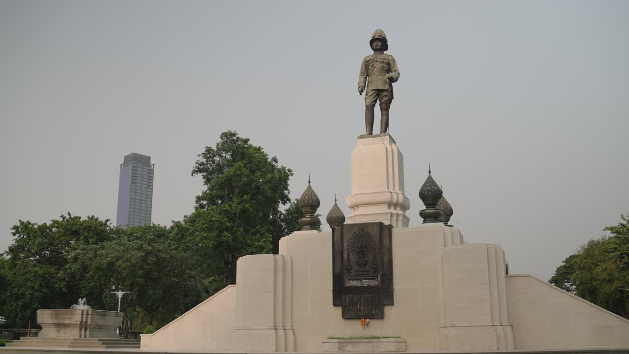 A medium, static shot of the iconic King Rama VI (Vajiravudh) monument at the entrance of Lumphini Park, Bangkok, Thailand, with the city skyline in the background