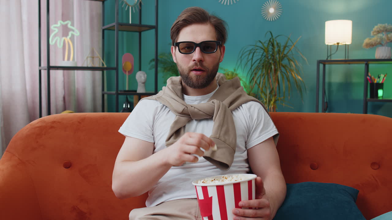Young man sits on couch eating popcorn and watching interesting tv serial sport game online at home