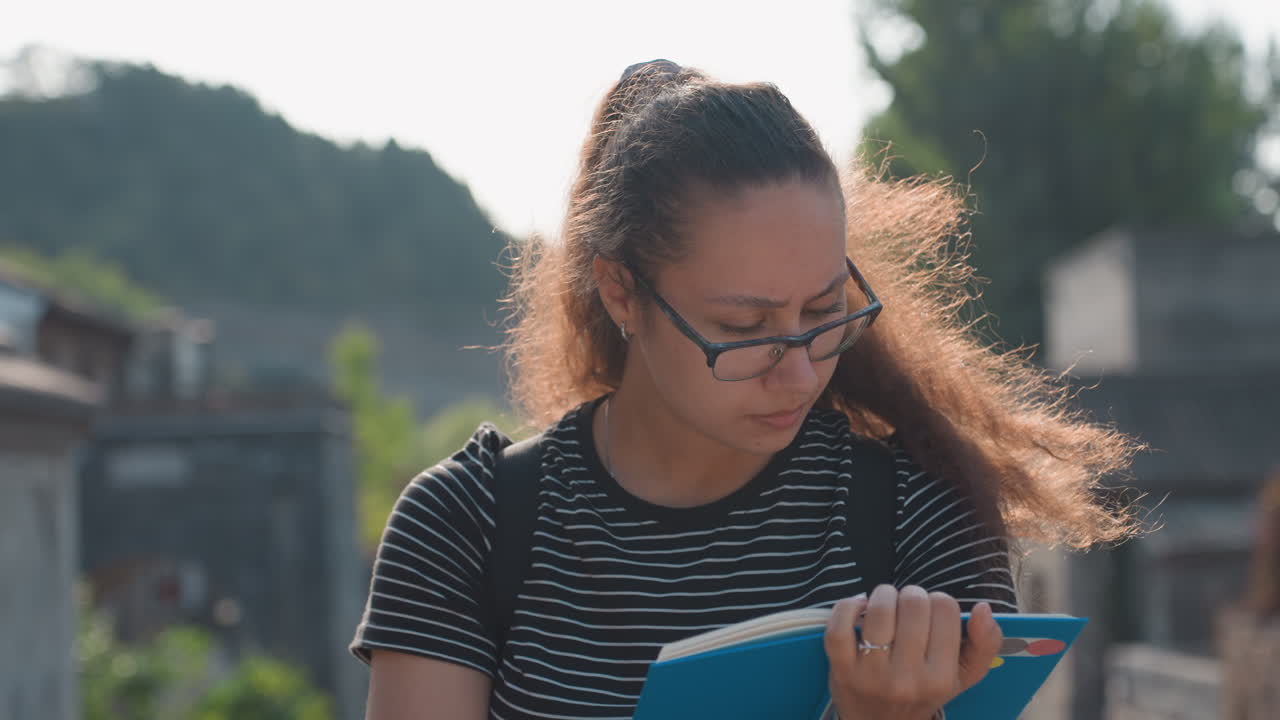 Woman sketches architecture in notebook on sunny street, focused student vibe with pen and glasses, heritage facades and quiet urban courtyard provide inspiration for study and art