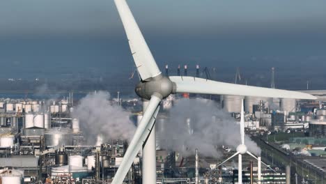 close up aerial orbiting shot of a wind turbine in a very dense industrial zone with stacks, steam and shiny steel tanks in the background on a sunny day near rotterdam netherlands