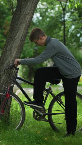 el niño camina hasta la bicicleta estacionada cerca del árbol en el campo cubierto de hierba, toca el pedal y coloca su pie en él mientras sostiene la bicicleta, en el fondo, el niño y la niña pasan en el fondo