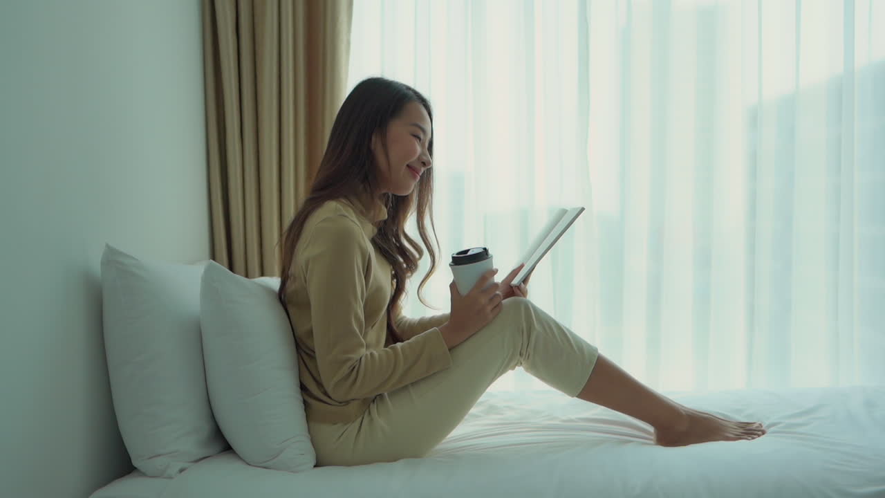 Young girl reading book, notebook, sitting on bed with coffee in hand