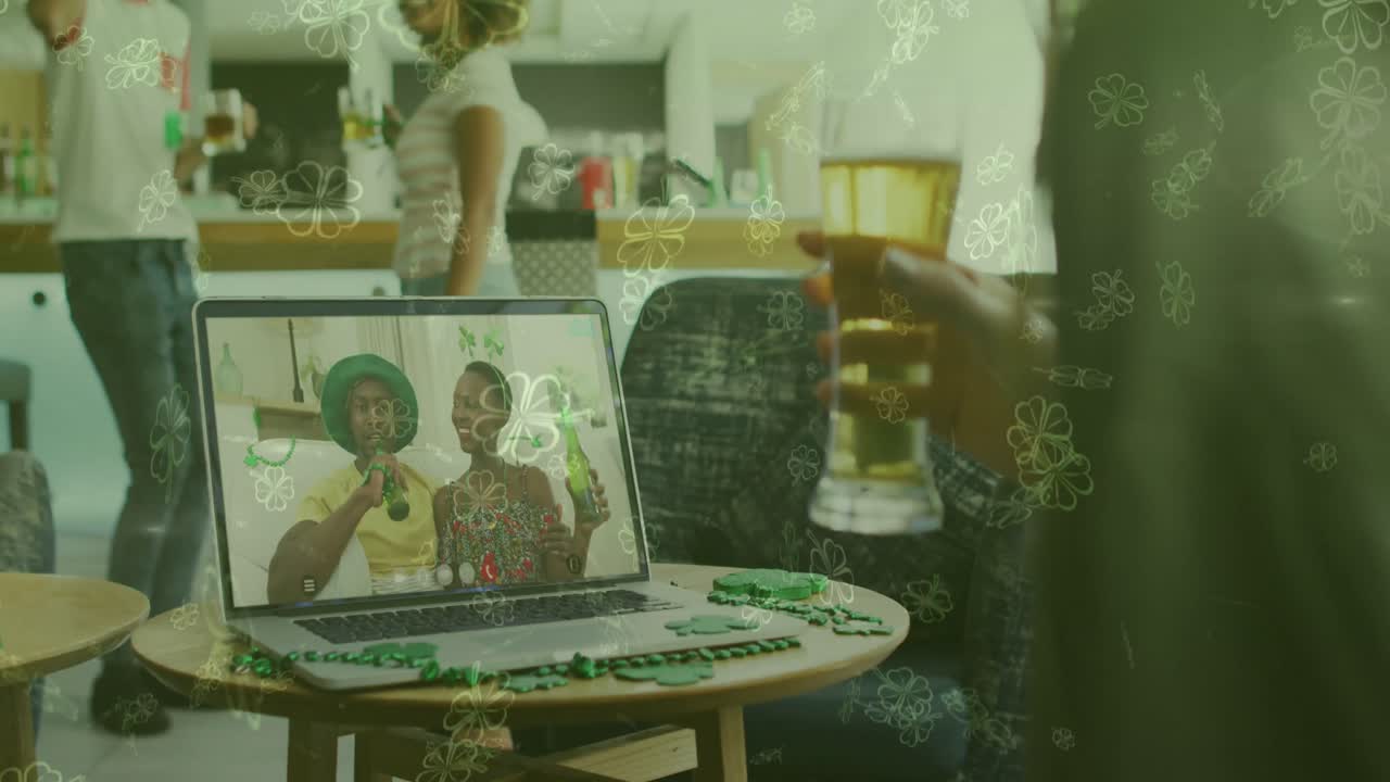 Animation of clover leaves over man celebrating st patrick's day with friends on laptop video call