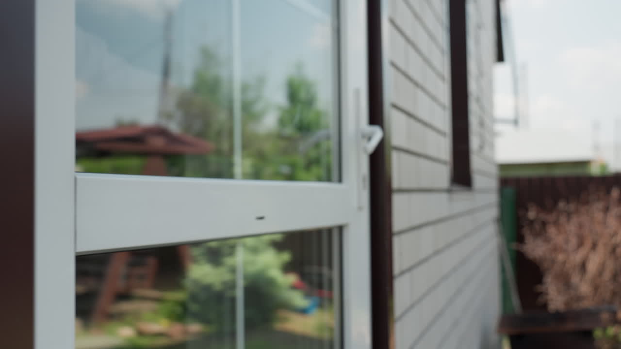 White Woman Entering Glass Patio Door, Suburban Exterior With Brick Wall, Towel Hanging On Hook, Glimpse Of Child Inside And Garden Planters, Casual Step Over Threshold, Reflective Window Panels