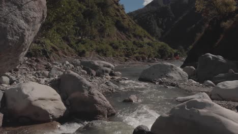 rocky-river-winding-through-mountains-in-canyon-valley-water-flowing-approaching-grey-boulders-green-trees-blue-sky-and-clouds-fast-aerial-moving-forward-proximity