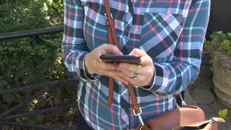 Eine-Frau-Mit-Kariertem-Hemd-Zieht-In-Einem-Park-Ihr-Handy-Aus-Einer-Braunen-Handtasche-Und-Schickt-Eine-Sms