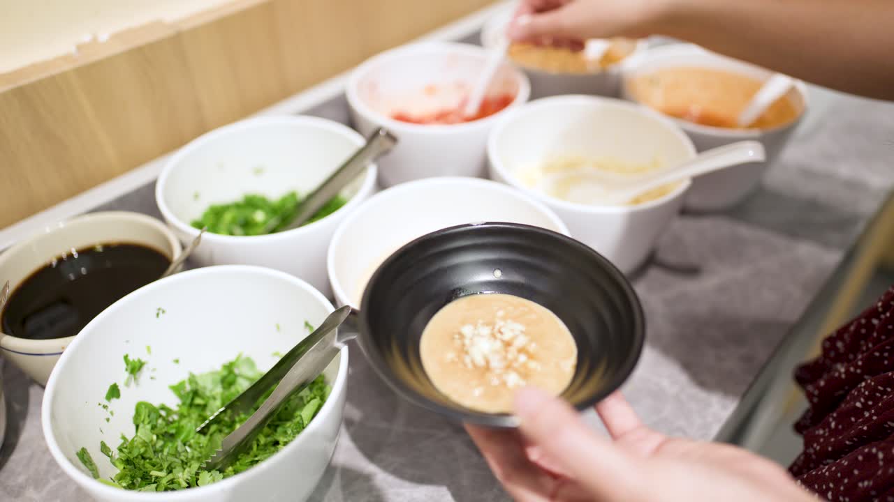 Hand adds chili to sesame sauce at a buffet condiment station, bright indoor lighting, close-up