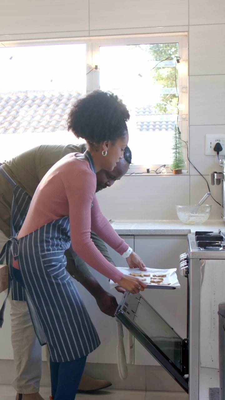 Vídeo vertical: Abrindo o forno, um casal de aventais em uma ilha de cozinha desliza uma assadeira para dentro do forno para assar.
