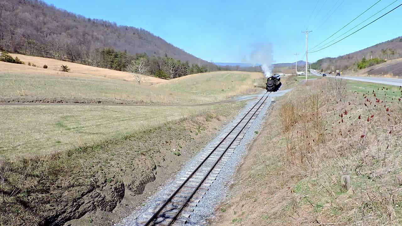 A historic steam train moves along a quiet railway in a beautiful countryside setting. It emits smoke while passing through rolling hills and greenery, under a clear blue sky