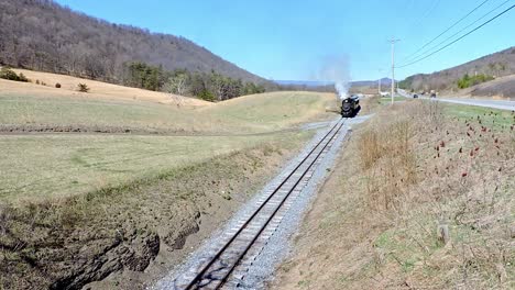 a historic steam train moves along a quiet railway in a beautiful countryside setting. it emits smoke while passing through rolling hills and greenery, under a clear blue sky